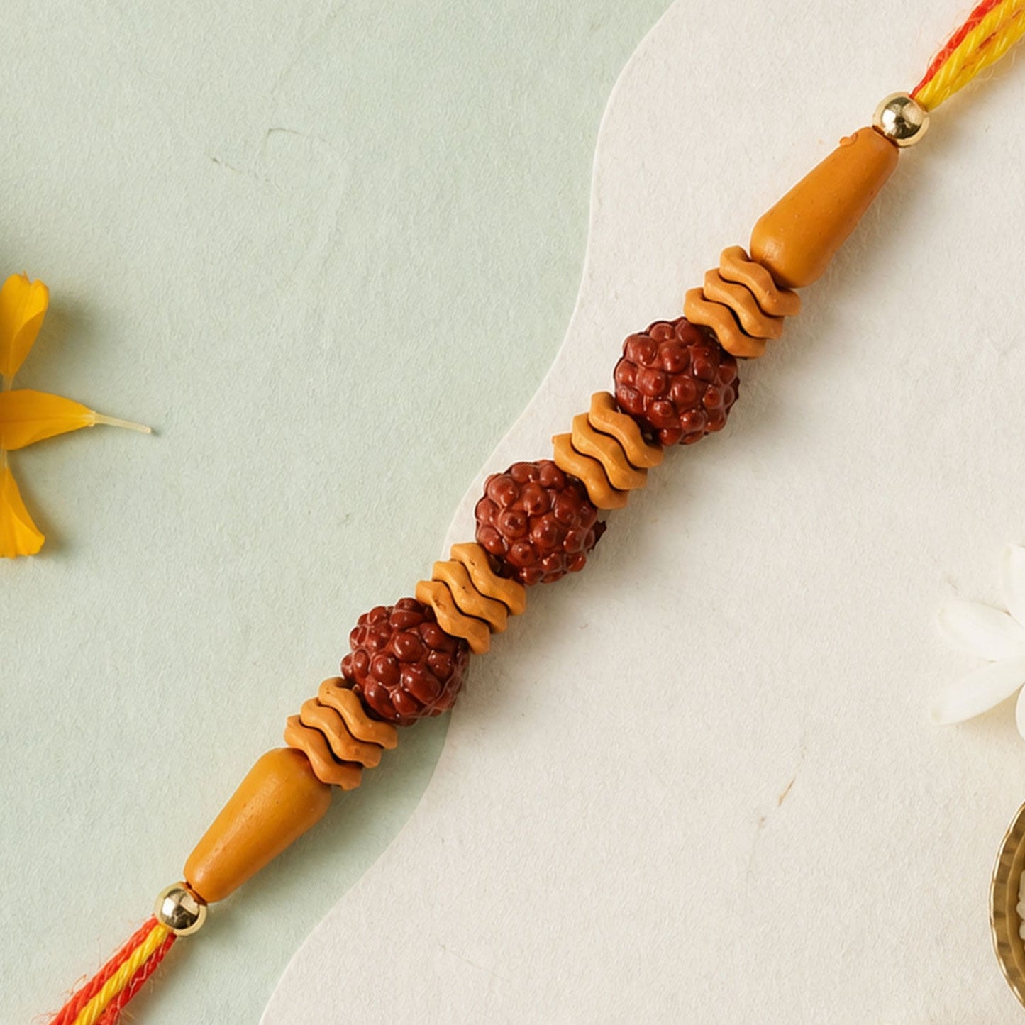 A Rudraksha Rakhi with three textured brown beads and wavy light brown spacers on a red and yellow thread, against a light