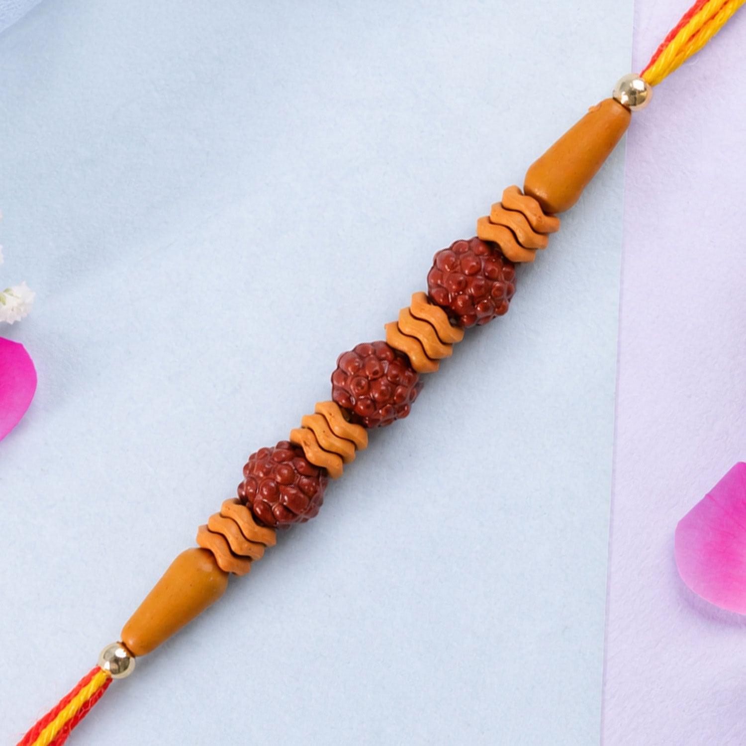 A Rudraksha Rakhi with three textured brown beads and wavy light brown spacers on a red and yellow thread, against  light