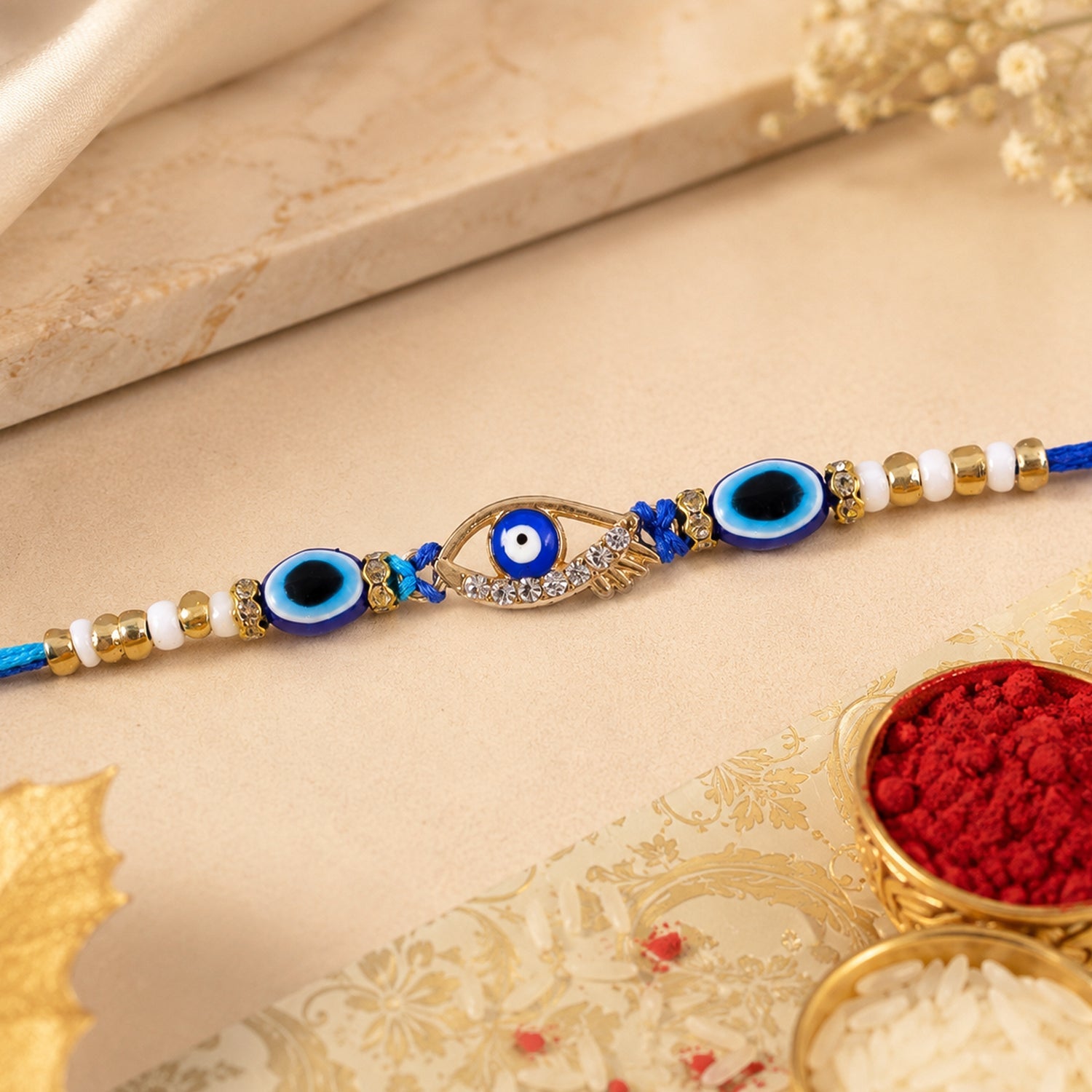 A blue evil eye rakhi with a gold and rhinestone center charm, flanked by smaller blue evil eye beads and white and gold