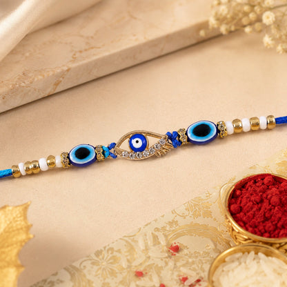 A blue evil eye rakhi with a gold and rhinestone center charm, flanked by smaller blue evil eye beads and white and gold