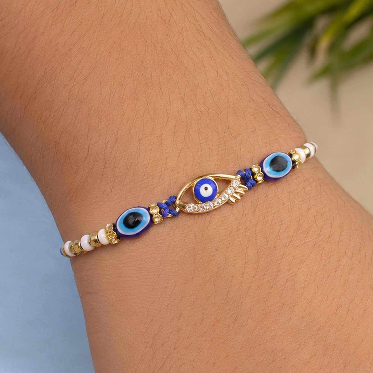 A blue evil eye rakhi with a gold and rhinestone center charm, featuring smaller blue evil eye beads and white and gold