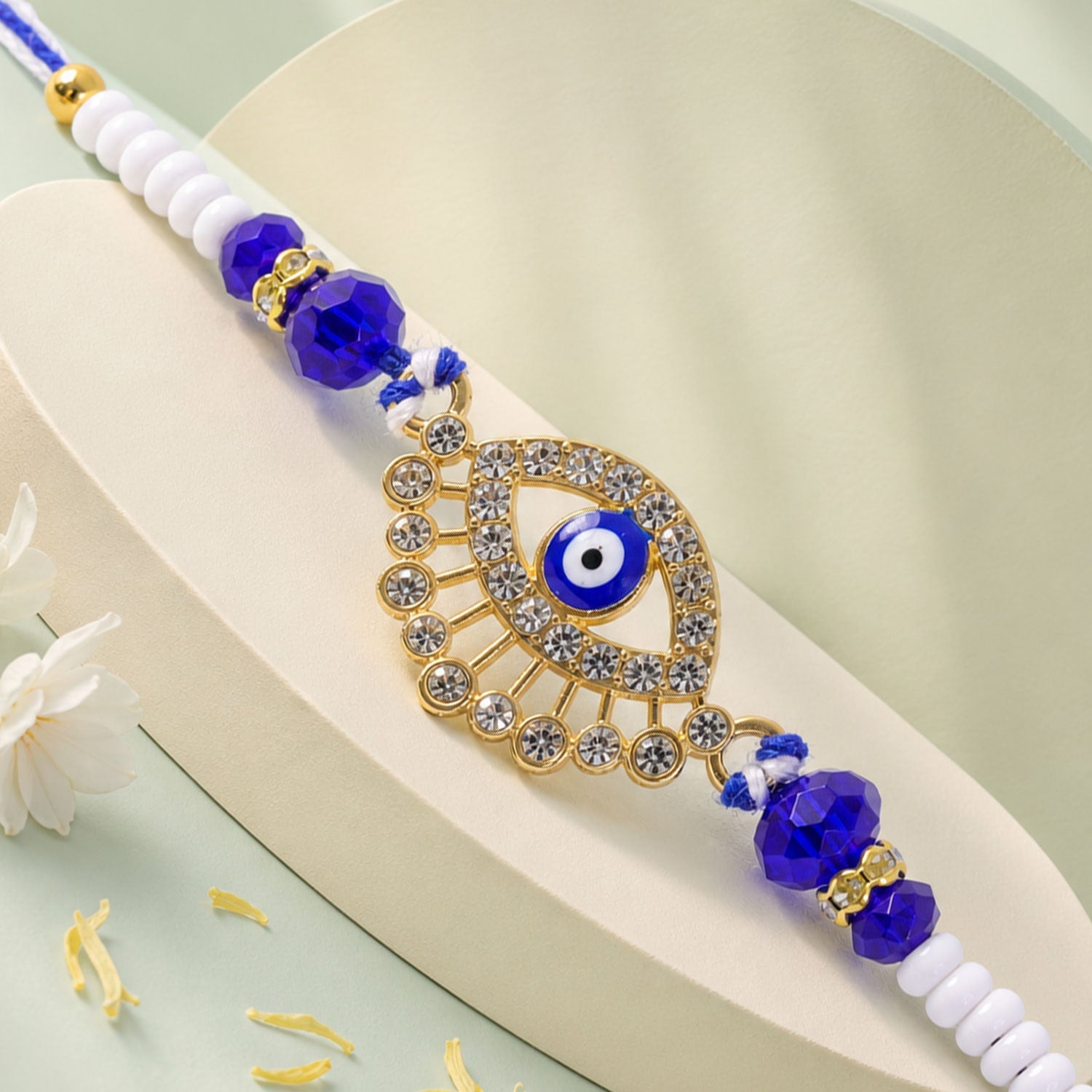 A gold and diamond evil eye Rakhi with blue and white beads on a white surface with white flowers.