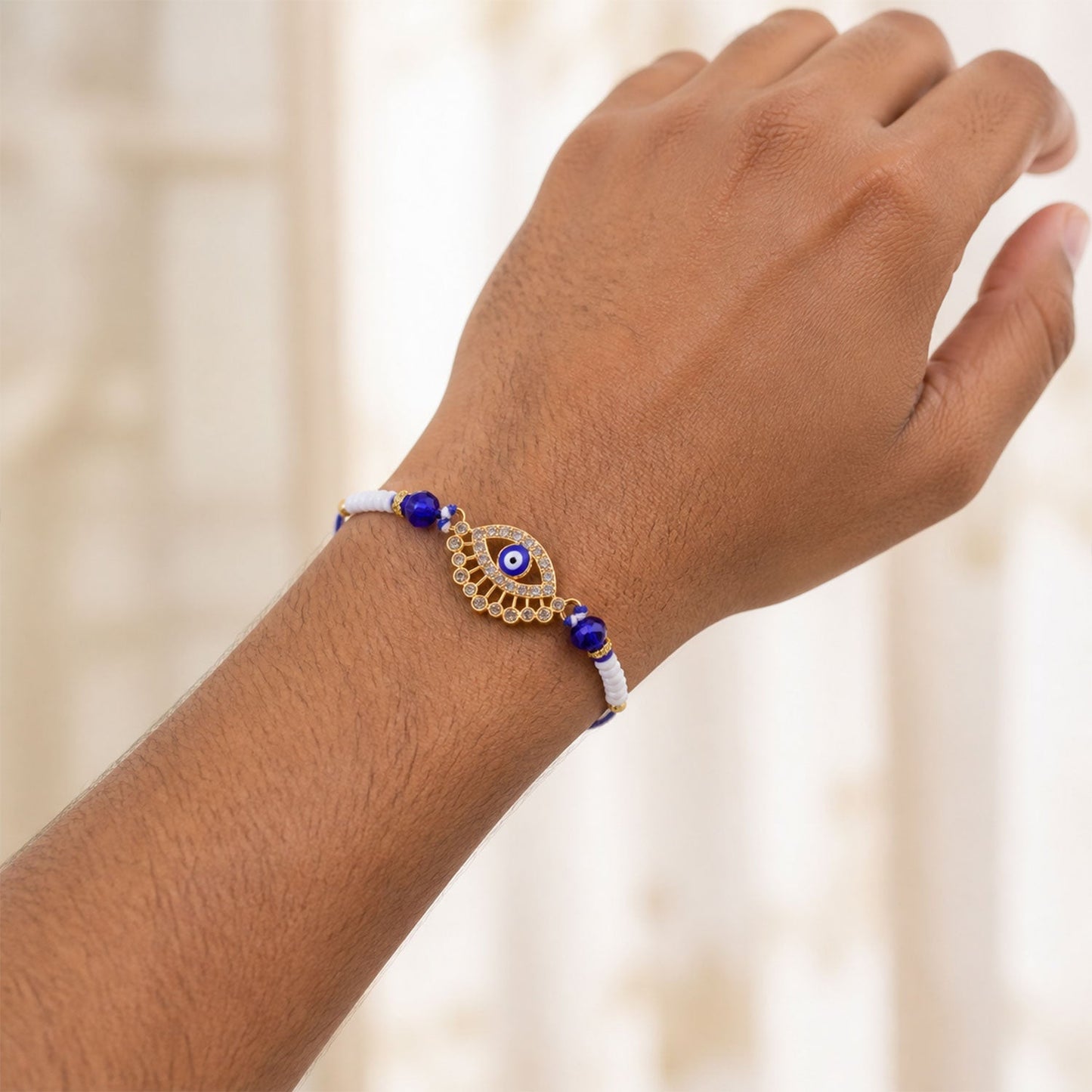 A person's arm wearing a gold and diamond evil eye Rakhi with blue and white beads.