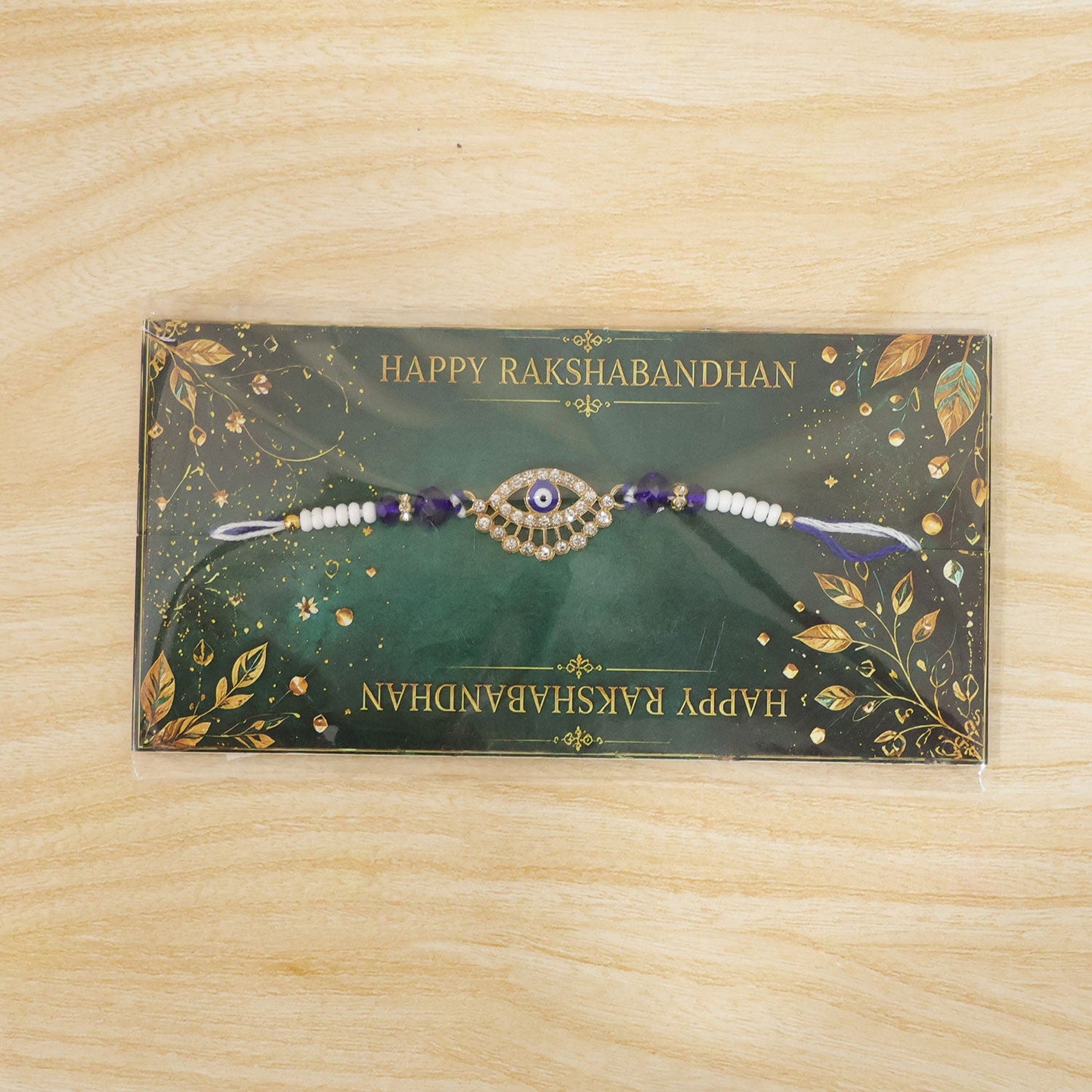 A gold and diamond evil eye Rakhi with blue and white beads on a green Happy Rakshabandhan card.