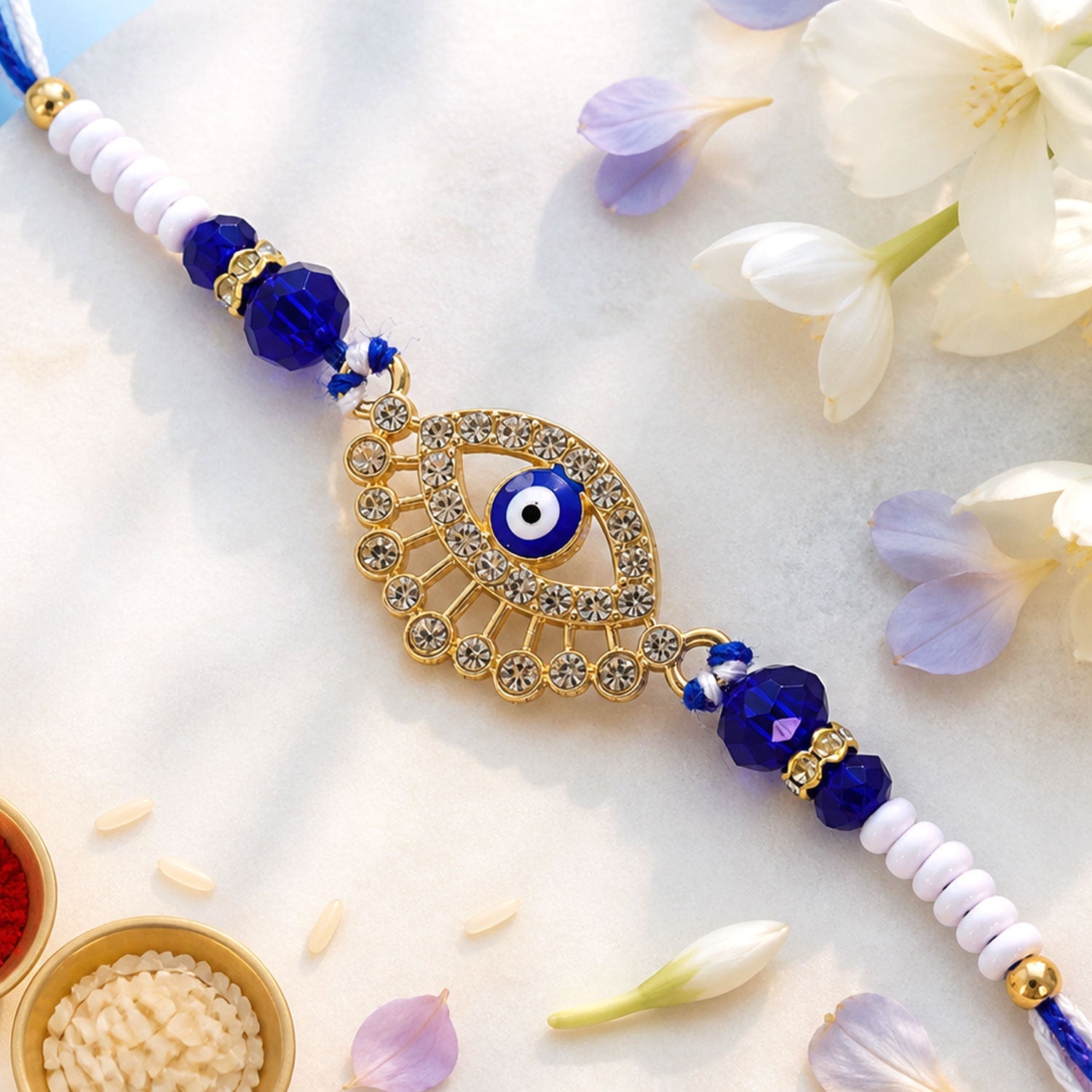 A gold and diamond evil eye Rakhi with blue and white beads on a white surface with rice and flowers.
