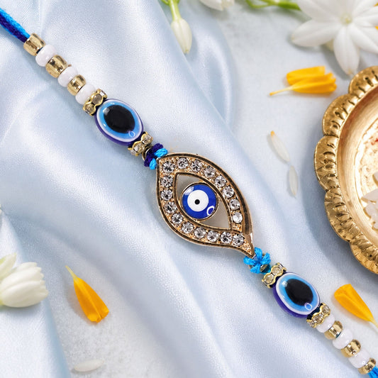 A blue evil eye rakhi with a stone-studded center charm on a light blue satin background with white flowers.