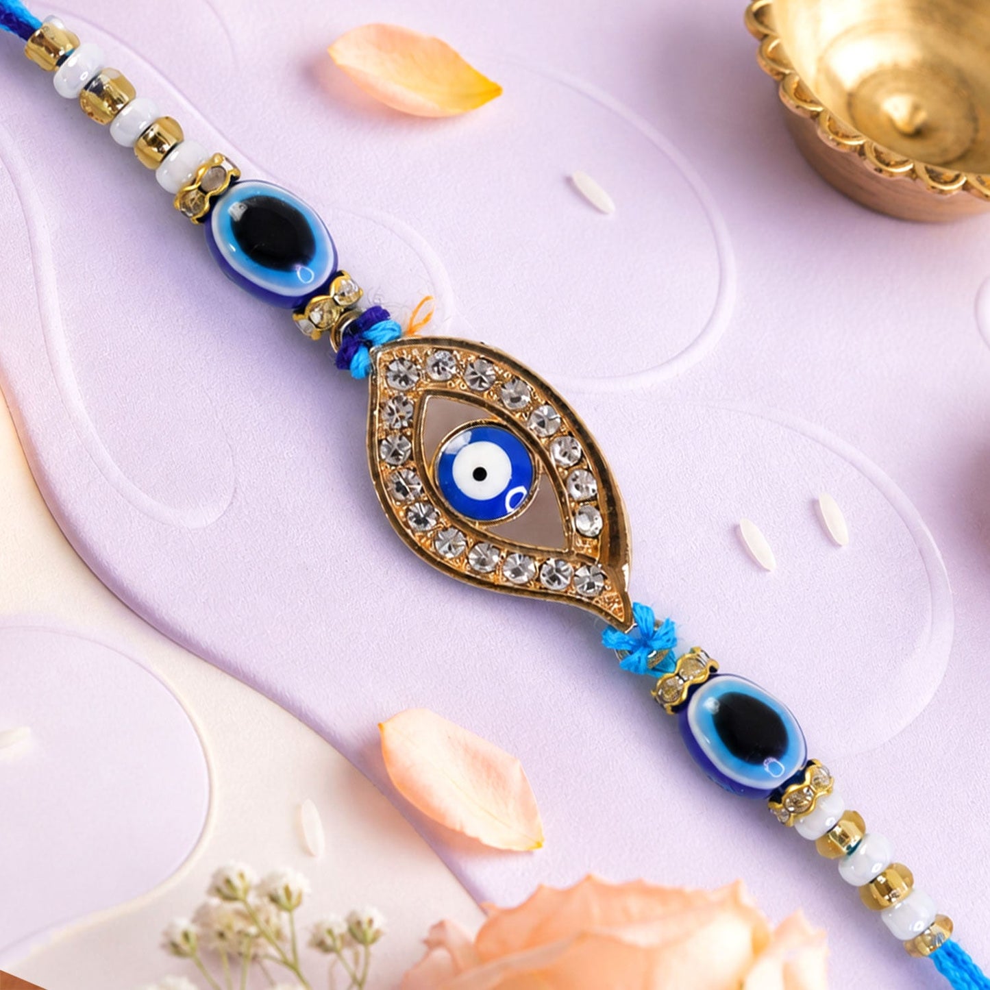 A blue evil eye rakhi with a stone-studded center charm on a light purple background with scattered rice grains.