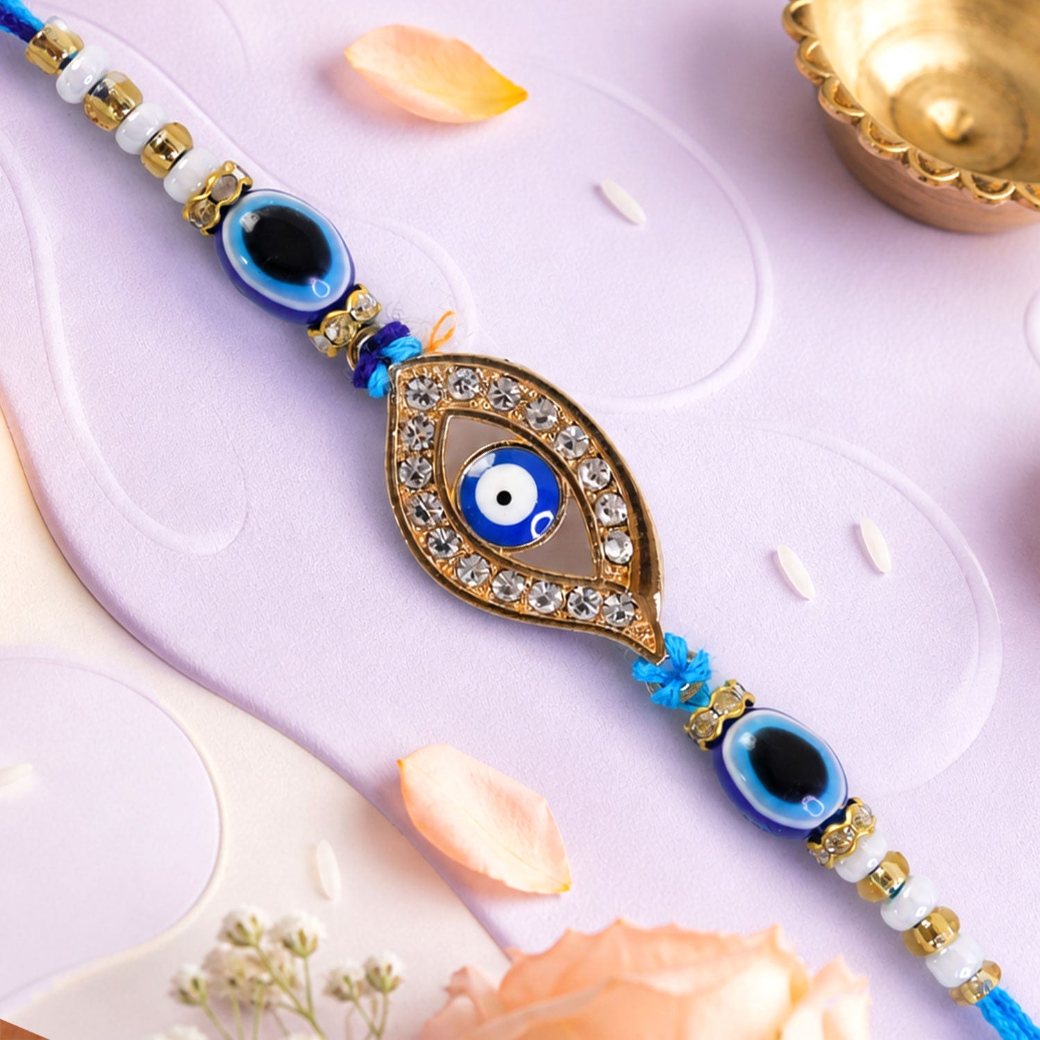 A blue evil eye rakhi with a stone-studded center charm on a light purple background with scattered rice grains.