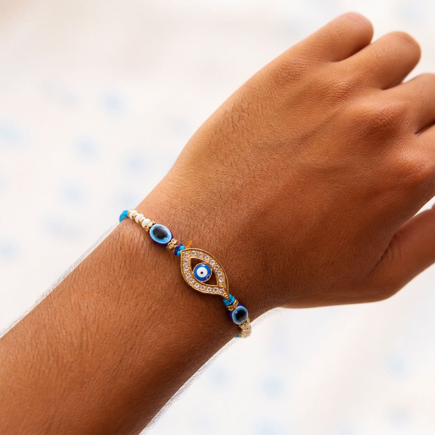 A blue evil eye rakhi with a stone-studded center charm on a person's wrist against a light background.