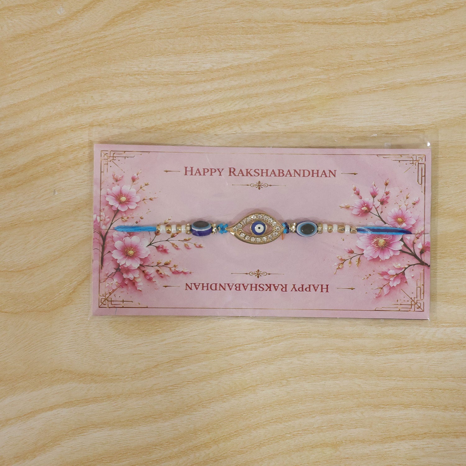 A blue evil eye rakhi on a pink 'Happy Rakshabandhan' card with cherry blossom designs, on a wooden surface.