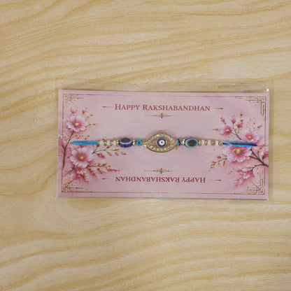 A blue evil eye rakhi on a pink 'Happy Rakshabandhan' card with cherry blossom designs, on a wooden surface.