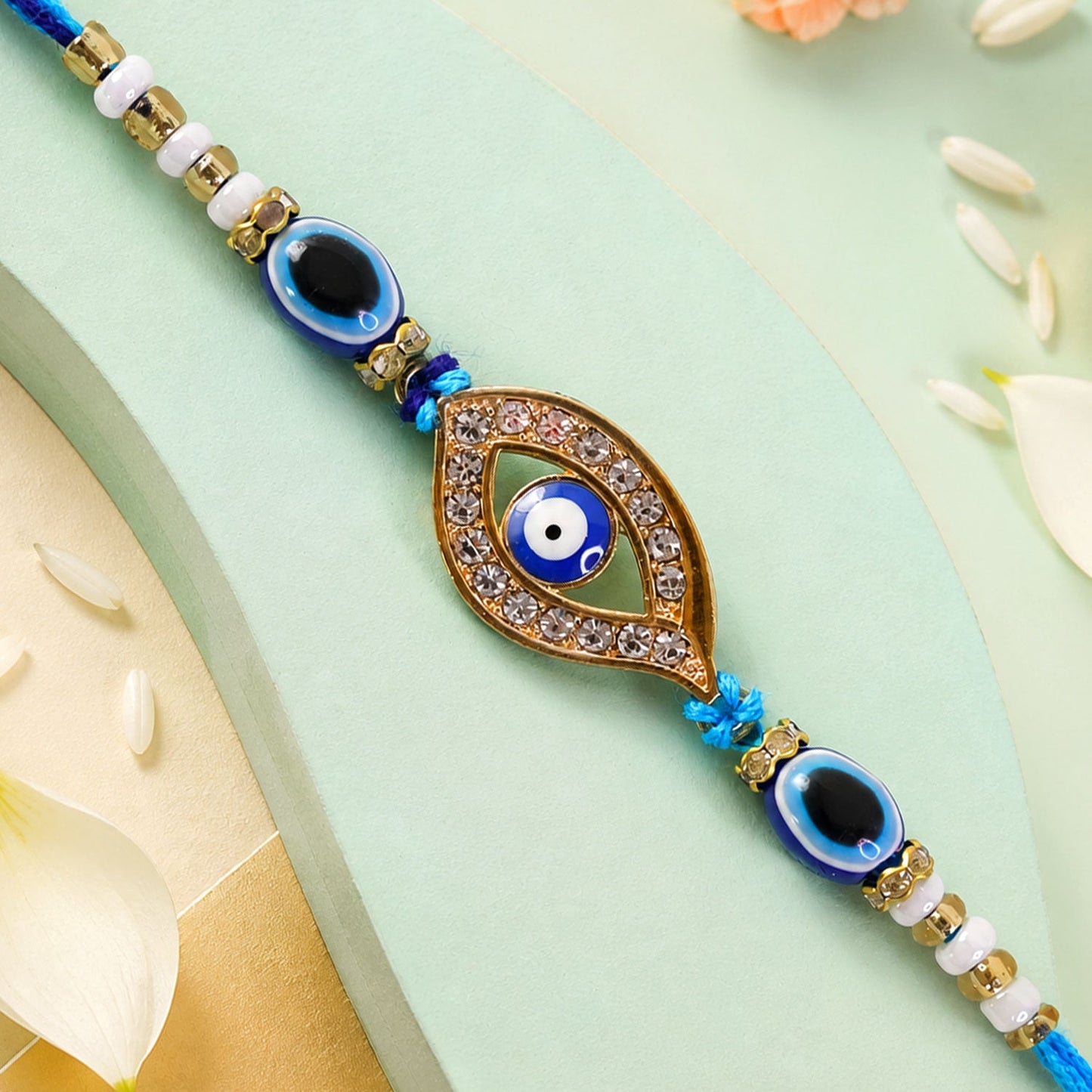 A blue evil eye rakhi with a stone-studded center charm on a light green background with white flower petals.