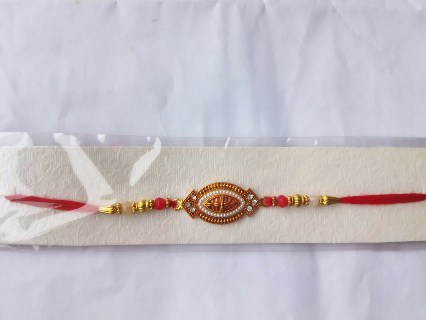A red and gold bracelet with an oval centerpiece, adorned with white beads and red stones, is displayed on a white textured