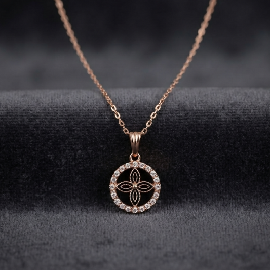 A rose gold necklace with a circular pendant featuring a floral design and clear stones, on a dark gray velvet surface.