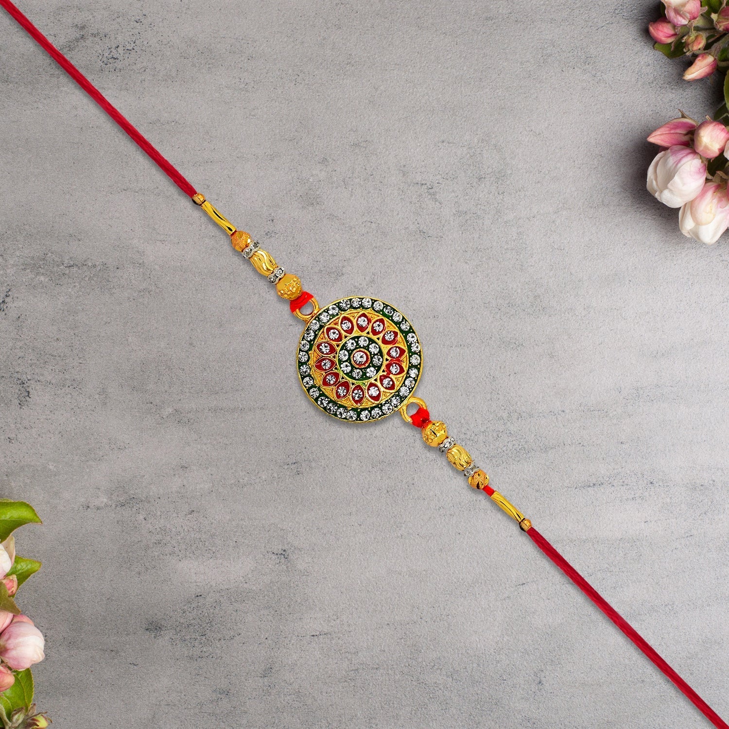 A red thread bracelet with a gold, green, and red circular medallion featuring clear gems, on a gray background with pink