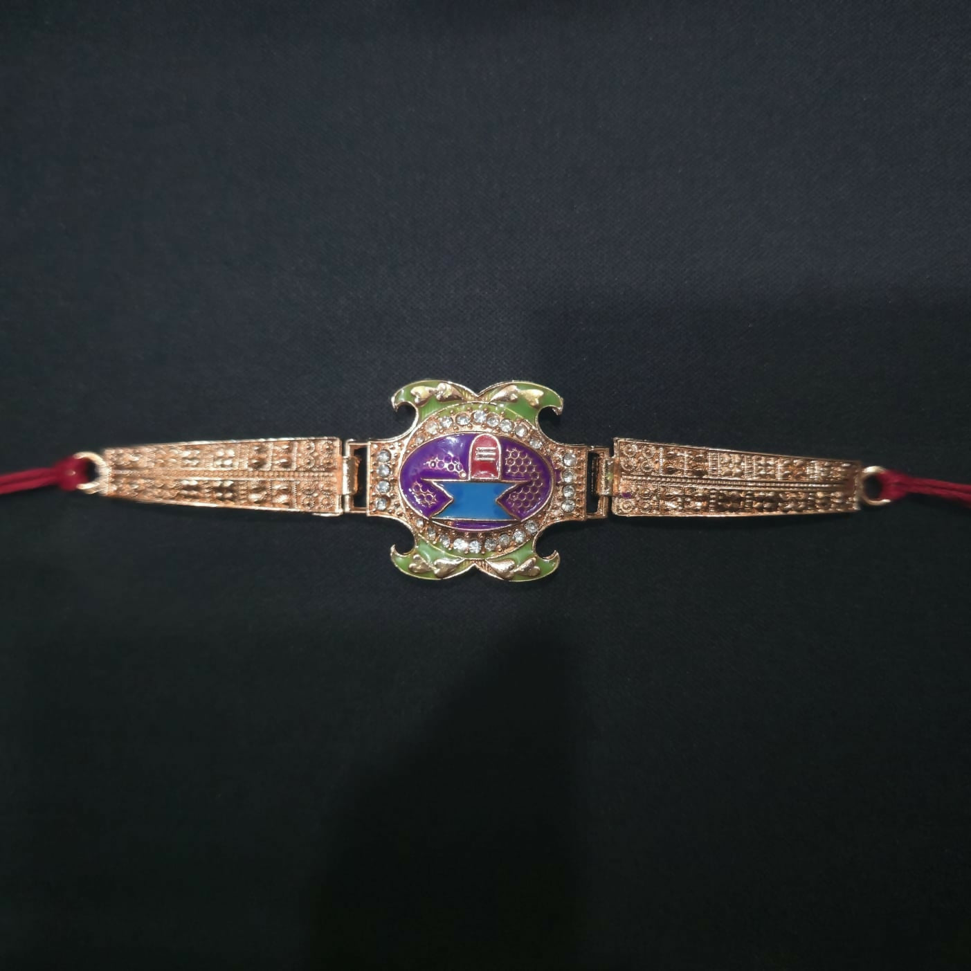 A rose gold Rakhi with a purple, blue, and green Shivling design is centered on a solid black background.