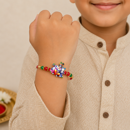 A child's arm, wearing a tan patterned shirt, displays a red thread with a colorful Bal Gopal Krishna and cow charm, flanked