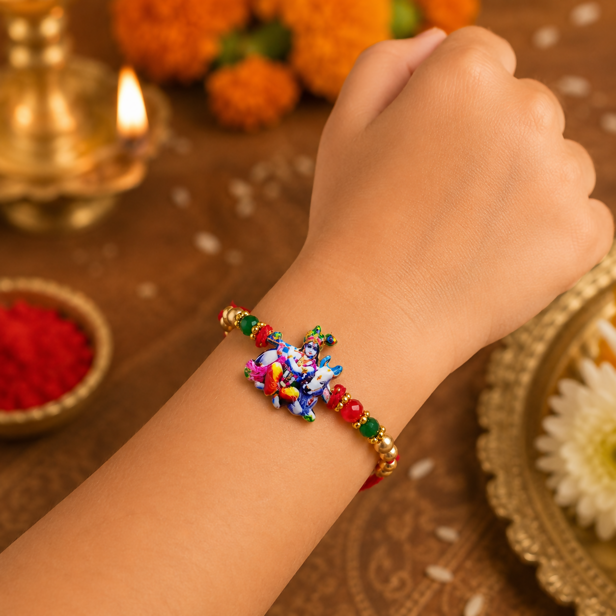 A child's arm wears a red thread with a colorful Bal Gopal Krishna and cow charm, flanked by red, green, and gold beads,