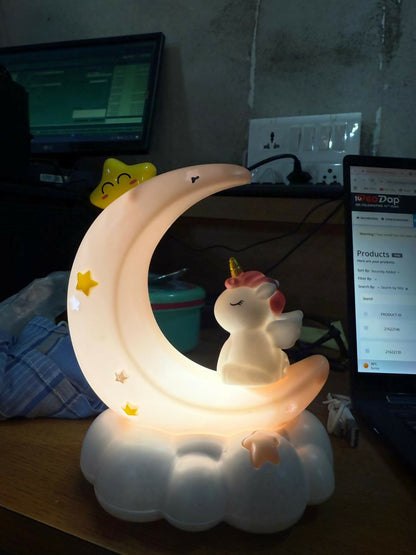 The glowing unicorn crescent moon night light sits on a desk next to a laptop in a dimly lit room.
