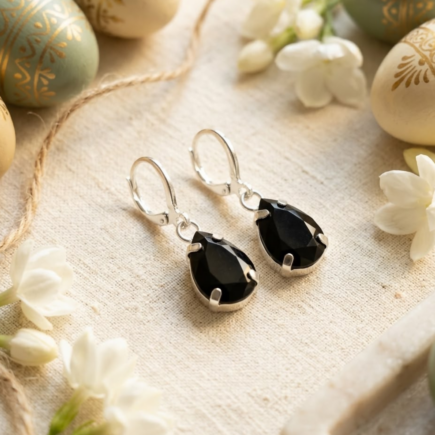 A pair of black teardrop crystal earrings on a white textured fabric, surrounded by white flowers and decorative eggs.
