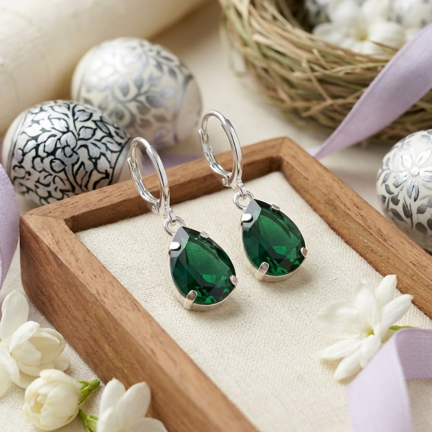 A pair of green teardrop crystal earrings with silver hooks rest on a wooden tray with decorative eggs and white flowers.