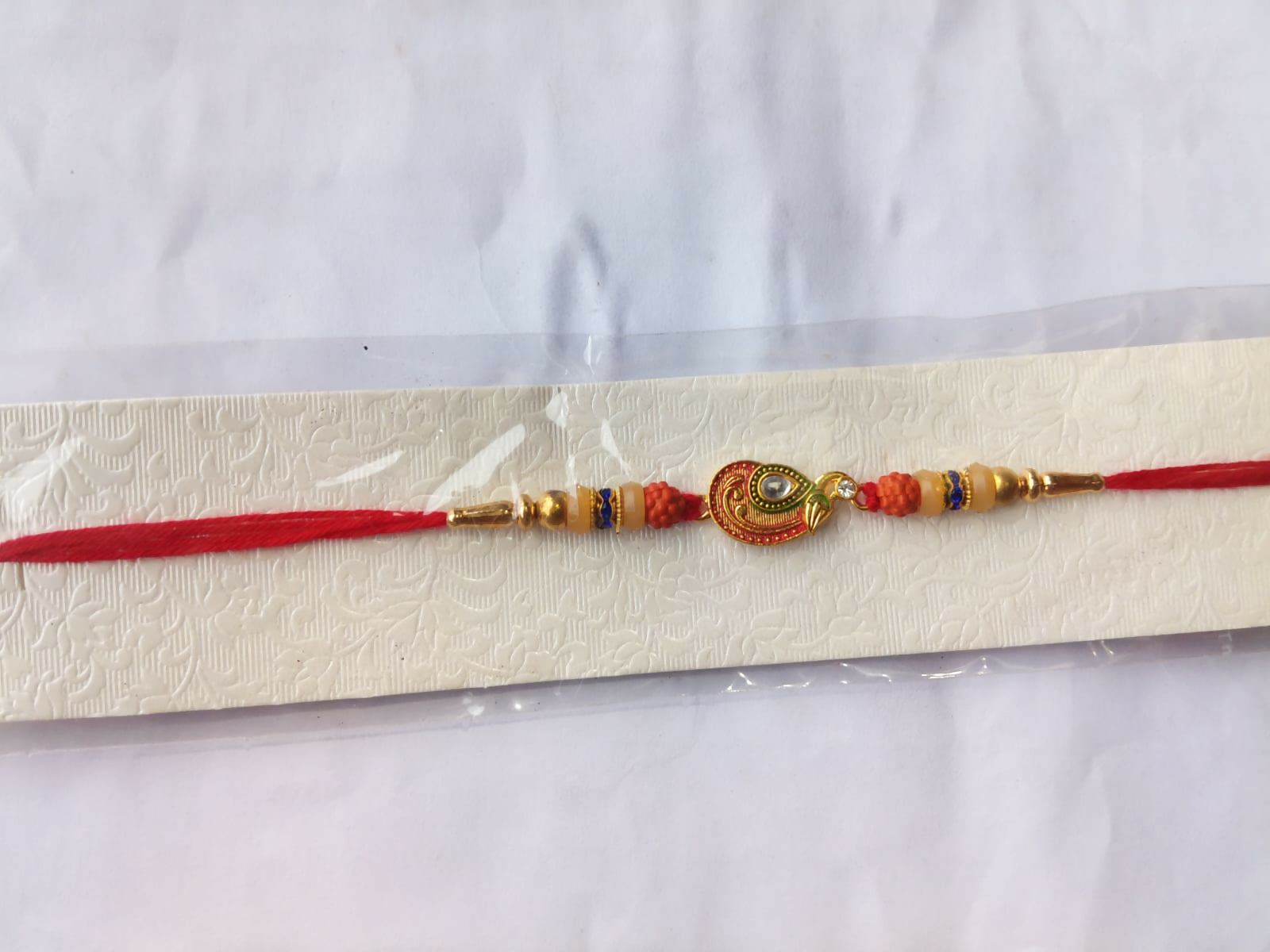 A red and gold bracelet with a peacock charm and orange and cream beads is displayed on a textured white card inside clear