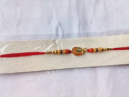 A red and gold bracelet with a peacock charm and orange and cream beads is displayed on a textured white card inside clear