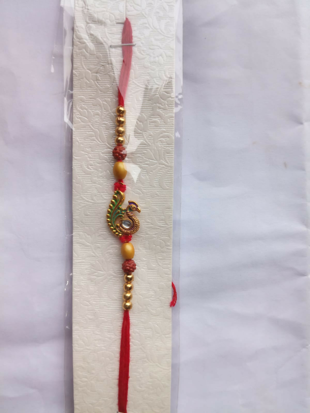 A peacock-decorated thread with gold and brown beads is displayed on a white, textured card, sealed in clear plastic