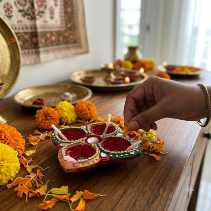 A hand places a wick into a red and green diya surrounded by marigolds and leaves on a wooden table.
