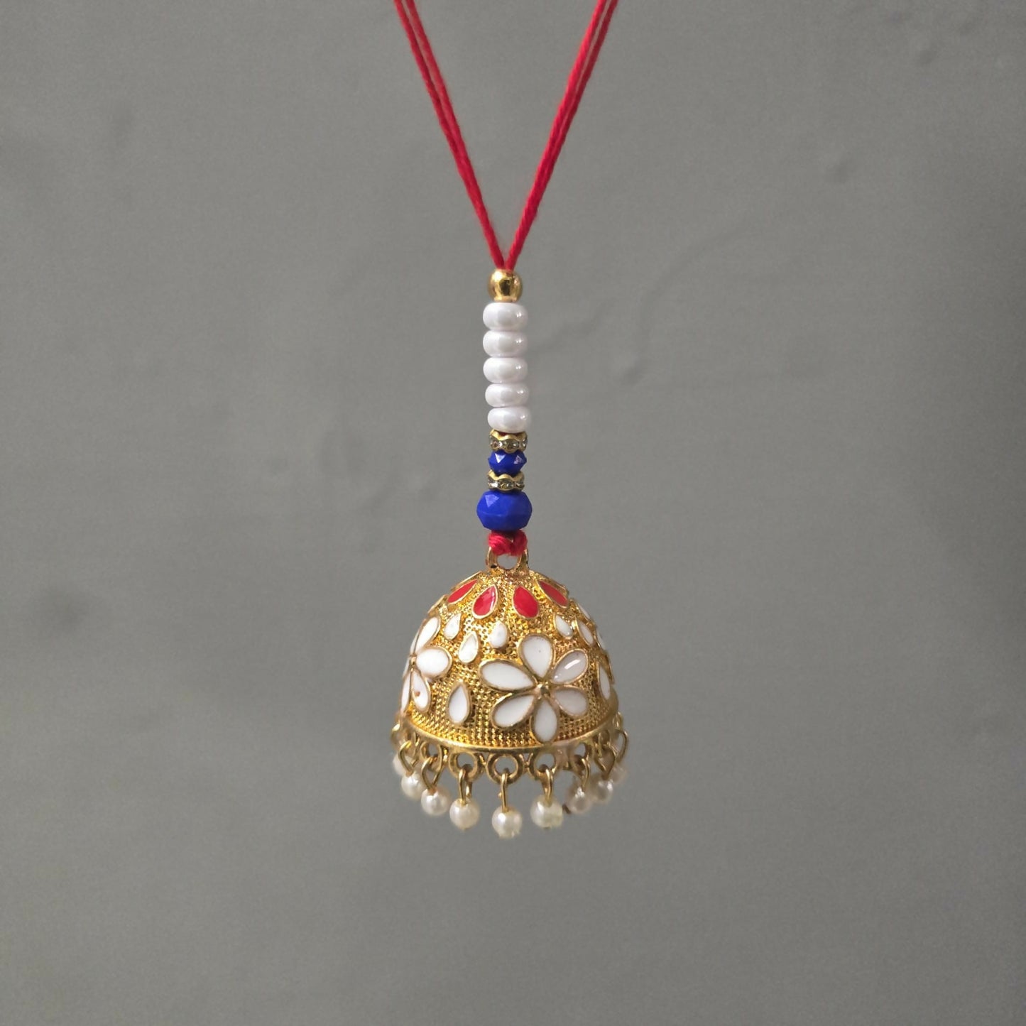 A gold-finished lumba with white and red enamel flowers, pearl drops, and a red thread hangs against a plain gray background.