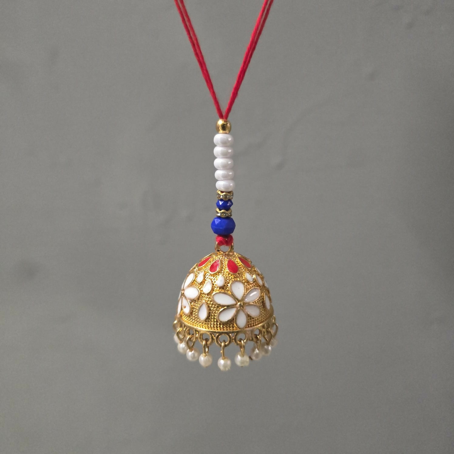 A gold-finished lumba with white and red enamel flowers, pearl drops, and a red thread hangs against a plain gray background.
