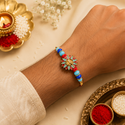 A man's arm wearing a red thread bracelet with a gold Ganesh charm, blue and light blue beads, and gold accents, with a diya
