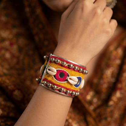 A person's arm wearing the colorful embroidered cuff bracelet, which features metal beads, mirrors, and shells, against a