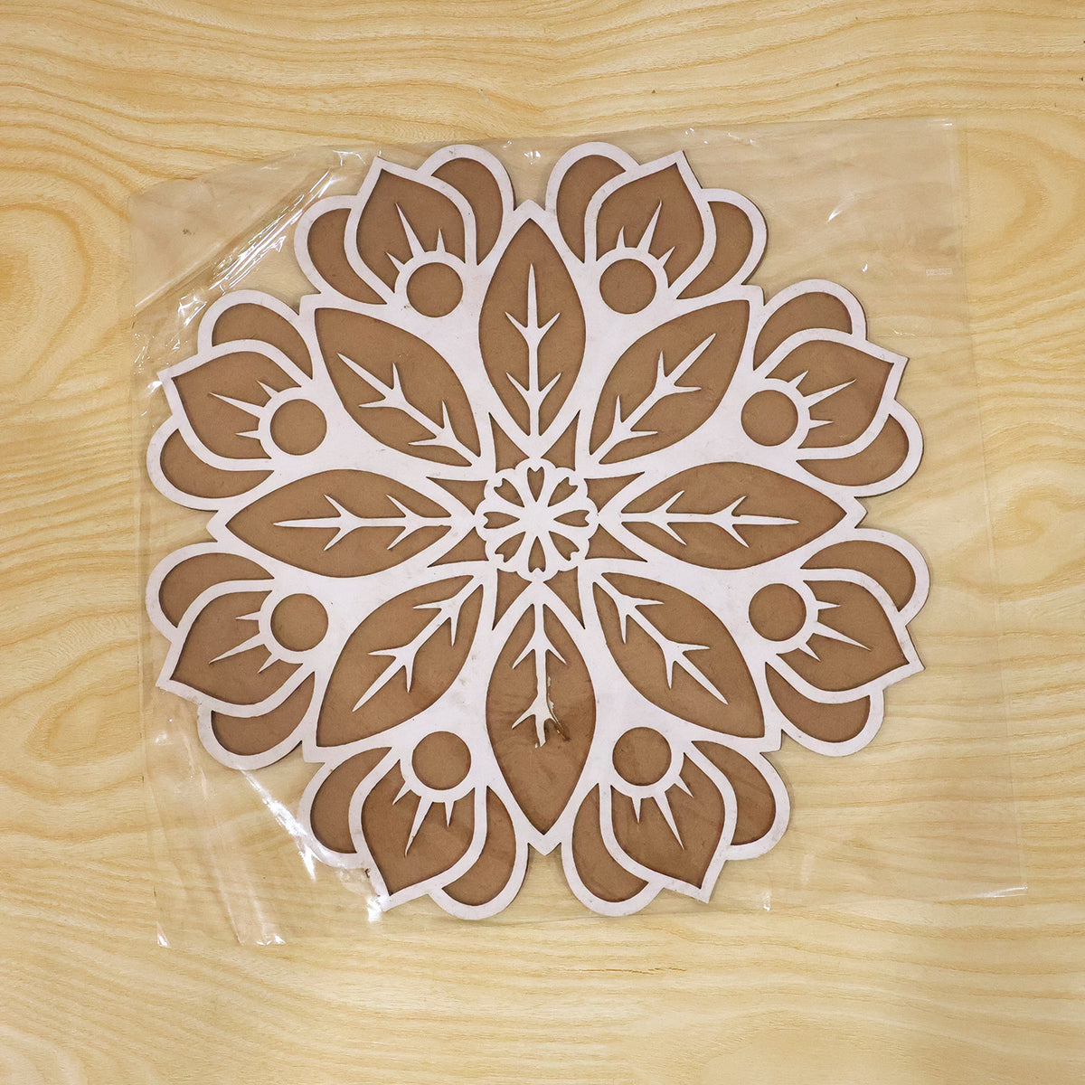 A white and brown wooden rangoli stencil is encased in clear plastic packaging, resting on a light wooden surface.