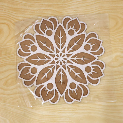 A white and brown wooden rangoli stencil is encased in clear plastic packaging, resting on a light wooden surface.