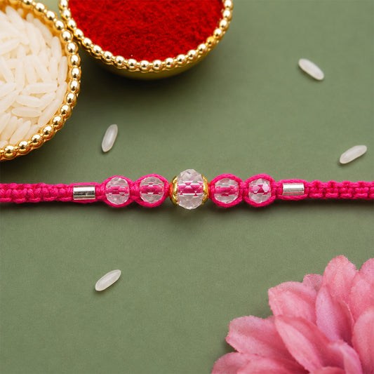 A pink beaded Rakhi with five clear crystal stones, a central gold-toned bead, and silver accents, on a green surface with