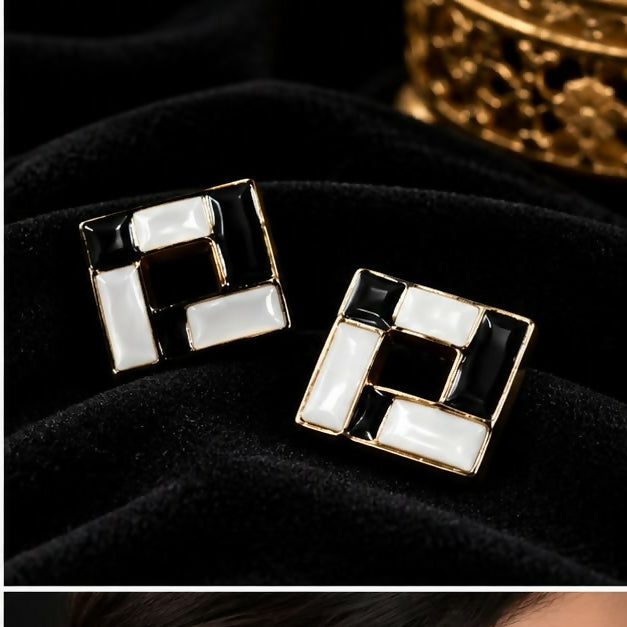Two black and white square earrings with gold borders rest on a black fabric.