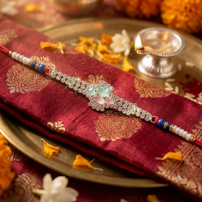 The elegant Rakhi, featuring a crystal floral design and blue beads, is displayed on a red silk cloth with gold patterns,