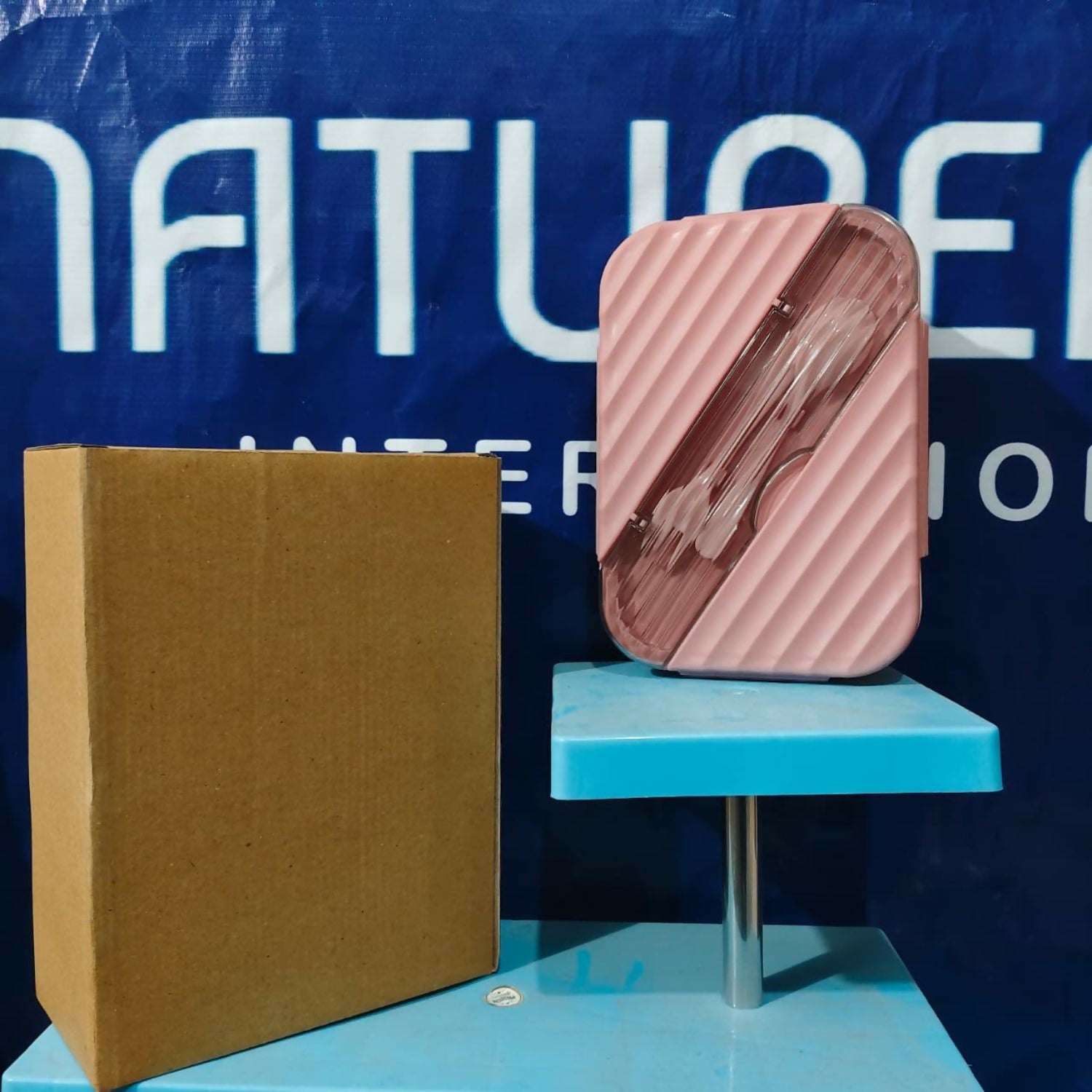 A pink bento lunch box with cutlery visible through its clear lid, standing upright next to a brown cardboard box.