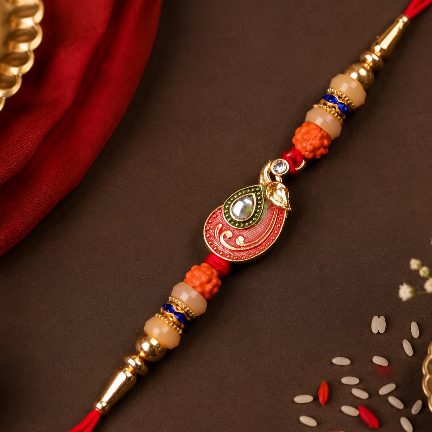 A red and gold bracelet with a peacock charm, orange and cream beads, and blue accents, rests on a dark brown surface with a