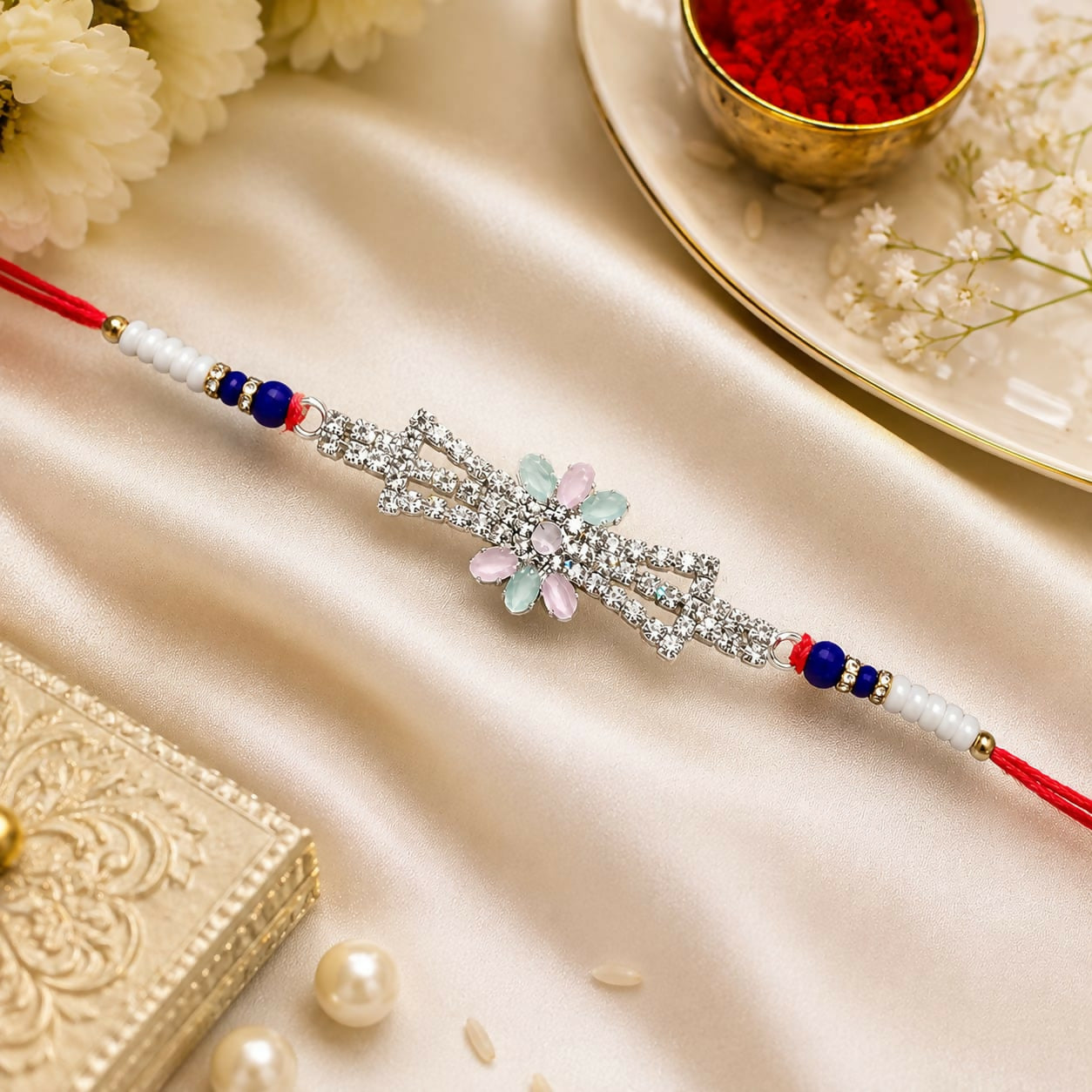A premium crystal Rakhi with blue beads and red thread lies on a light cream satin fabric beside a decorative box and pearls.